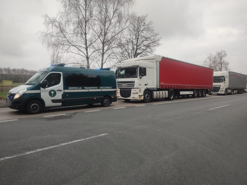 Na zdjęciu widoczny jest samochód INSPEKCJI TRANSPORTU DROGOWEGO’ oraz 2 duże ciężarówki zaparkowane na poboczu drogi. W tle widać szare niebo i bezlistne drzewa, co sugeruje pochmurny dzie