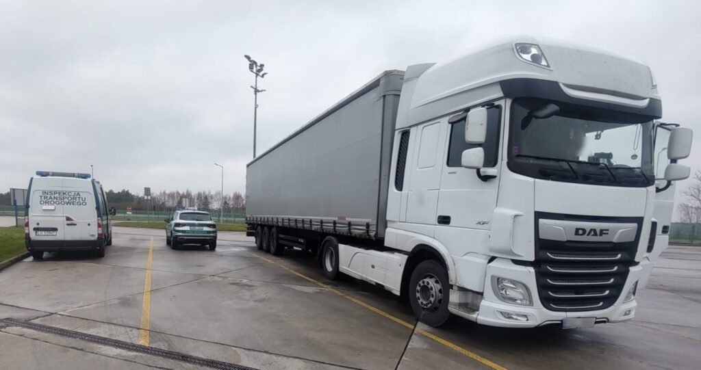 Biały ciągnik siodłowy z naczepą zaparkowany na asfaltowym parkingu. Po lewej stronie znajduje się samochód ITD z częściowo widocznym napisem. W tle widać bezlistne drzewa i szare niebo, sugerujące pochmurny dzień.