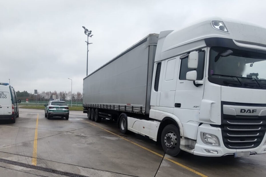 Biały ciągnik siodłowy z naczepą zaparkowany na asfaltowym parkingu. Po lewej stronie znajduje się samochód ITD z częściowo widocznym napisem. W tle widać bezlistne drzewa i szare niebo, sugerujące pochmurny dzień.