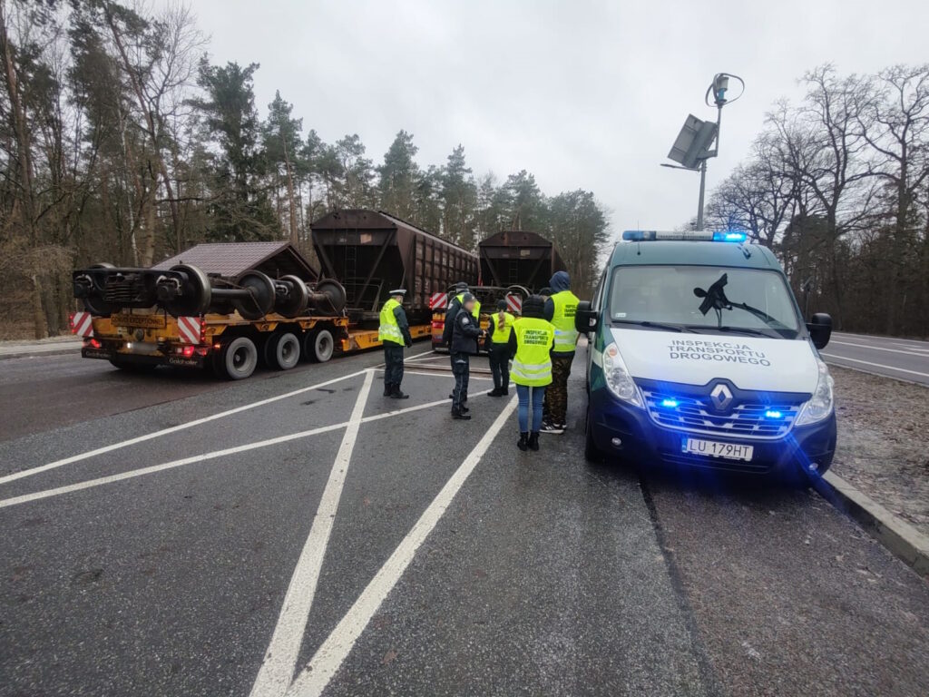 Inspektorzy transportu drogowego w odblaskowych kamizelkach stoją obok niebieskiego furgonu z włączonymi światłami sygnalizacyjnymi, na parkingu przy drodze. Obok znajduje się naczepa przewożąca wagon kolejowy.