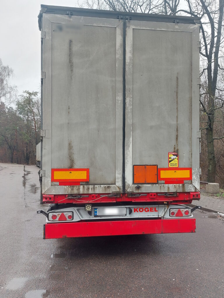 Zdjęcie przedstawia tył naczepy ciężarowej marki Kögel, zaparkowanej na poboczu drogi. Naczepa ma szare drzwi z widocznymi śladami zużycia i zabrudzenia. Na drzwiach znajdują się pomarańczowe i czerwone odblaski, a po prawej stronie naklejka ostrzegawcza z napisem "Attention Angles Mortes". Tablica rejestracyjna jest zamazana. Tło zdjęcia stanowią drzewa bez liści oraz chodnik.