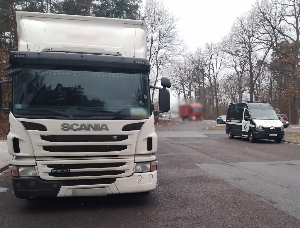 Biały ciągnik siodłowy marki Scania P230 zaparkowany na placu kontrolnym. W tle widoczny jest pojazd Inspekcji Transportu Drogowego (ITD) oraz inne samochody na drodze, otoczone zimową scenerią z drzewami bez liści.