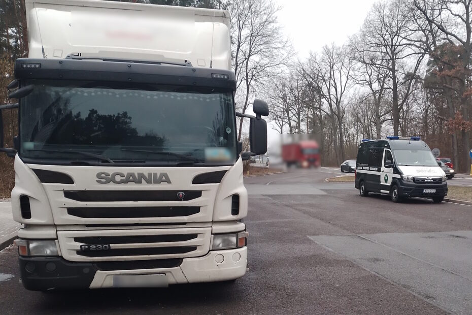 Biały ciągnik siodłowy marki Scania P230 zaparkowany na placu kontrolnym. W tle widoczny jest pojazd Inspekcji Transportu Drogowego (ITD) oraz inne samochody na drodze, otoczone zimową scenerią z drzewami bez liści.