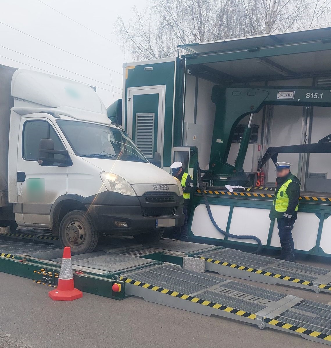 Biały samochód dostawczy marki Iveco stoi na mobilnej wadze kontrolnej, obsługiwanej przez inspektorów w odblaskowych kamizelkach. W tle widoczny jest specjalistyczny pojazd wyposażony w sprzęt do ważenia pojazdów.