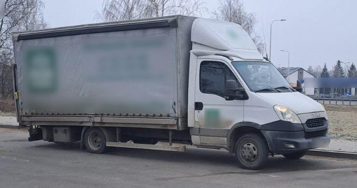 Biały samochód dostawczy marki Iveco z plandeką zaparkowany na poboczu drogi. W tle widoczna jest zimowa sceneria z drzewami i budynkami.