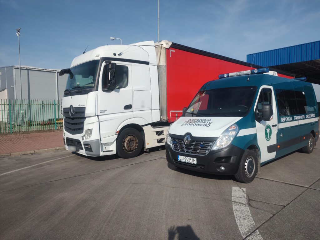 "Ciężarówka marki Mercedes Actros oraz pojazd Inspekcji Transportu Drogowego marki Renault Master stoją obok siebie na parkingu, w tle widoczne ogrodzenie i budynki przemysłowe."