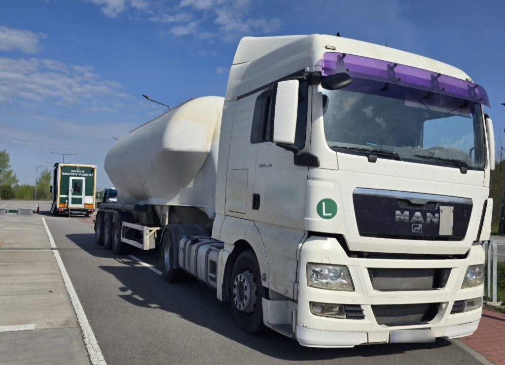 Biała ciężarówka MAN z naczepą typu cysterna stoi zaparkowana na parkingu przy drodze, w tle widoczny jest pojazd inspekcji transportu drogowego.