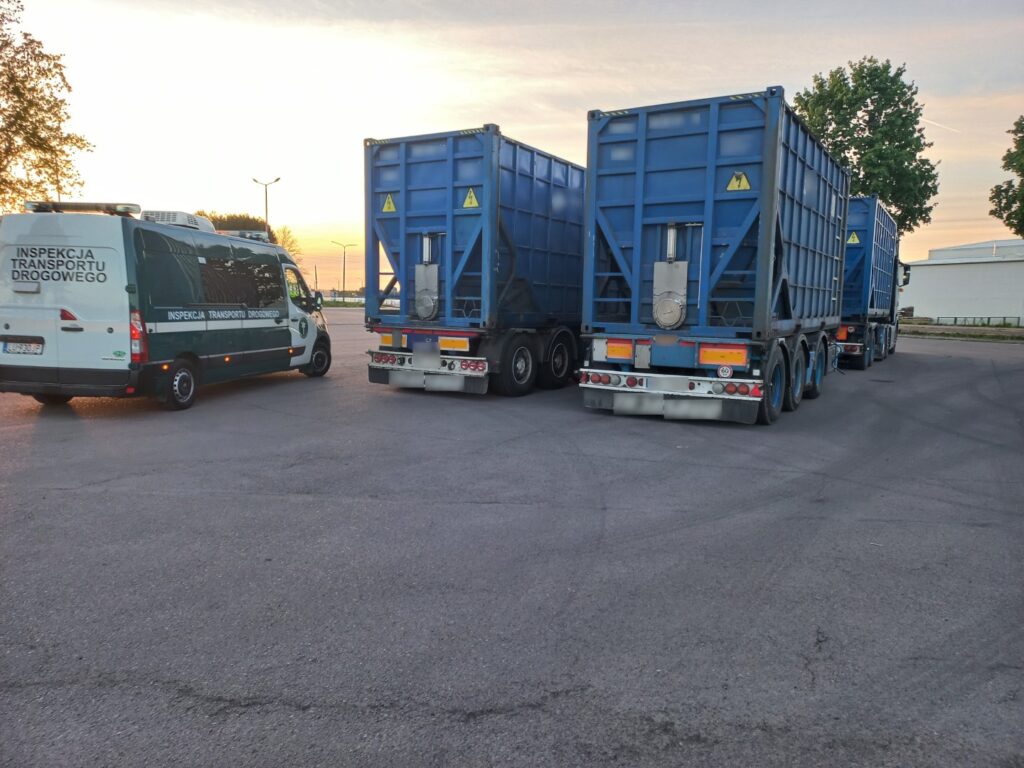 Na zdjęciu widoczne są dwa niebieskie samochody ciężarowe oraz biały pojazd Inspekcji Transportu Drogowego zaparkowane na placu asfaltowym o zachodzie słońca.