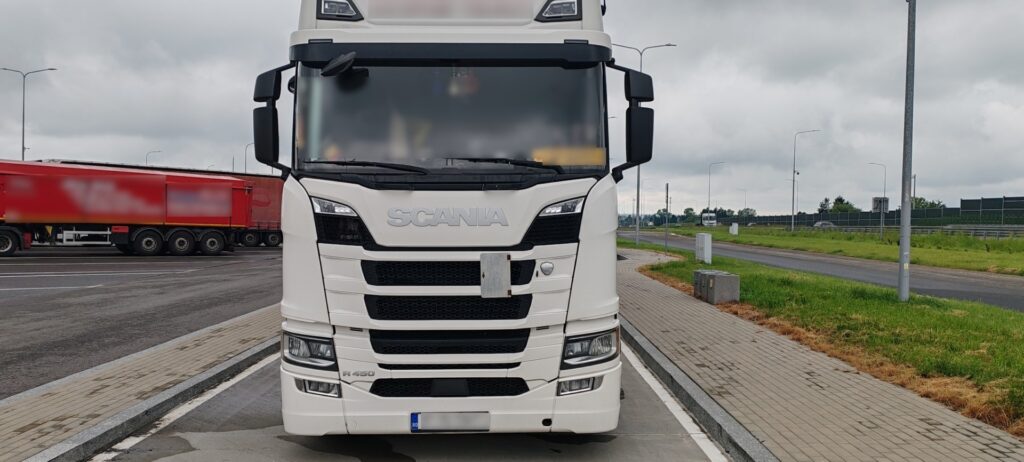 Przód białej ciężarówki marki Scania zaparkowanej na parkingu przy drodze, widoczny numer rejestracyjny częściowo zamazany.