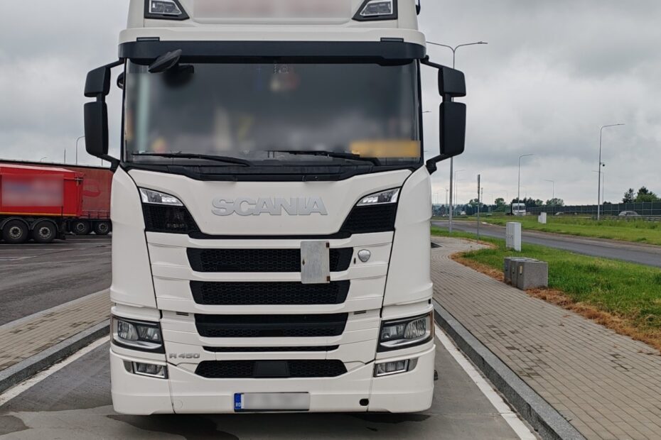 Przód białej ciężarówki marki Scania zaparkowanej na parkingu przy drodze, widoczny numer rejestracyjny częściowo zamazany.
