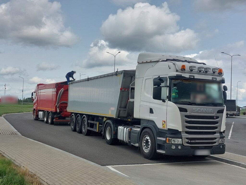 Biała ciężarówka Scania z naczepą, obok czerwony samochód ciężarowy i osoba stojąca na naczepie