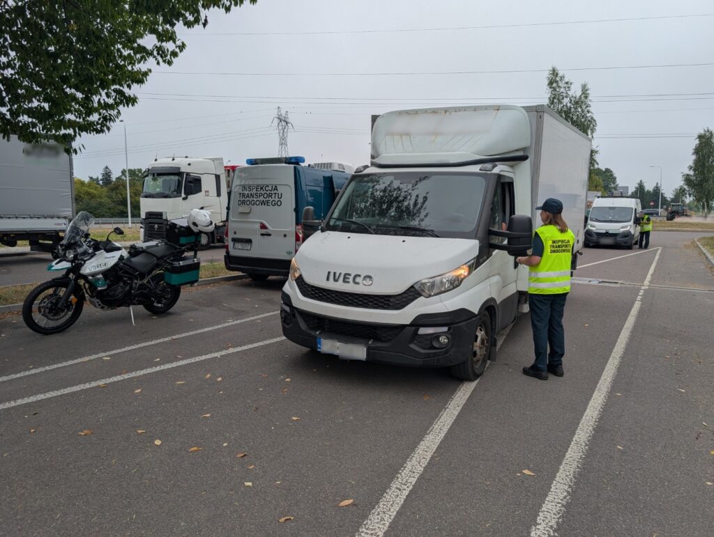 Kontrola drogowa prowadzona przez Inspekcję Transportu Drogowego; inspektor rozmawia z kierowcą dostawczego Iveco na parkingu obok radiowozu, motocykla służbowego i innych pojazdów.