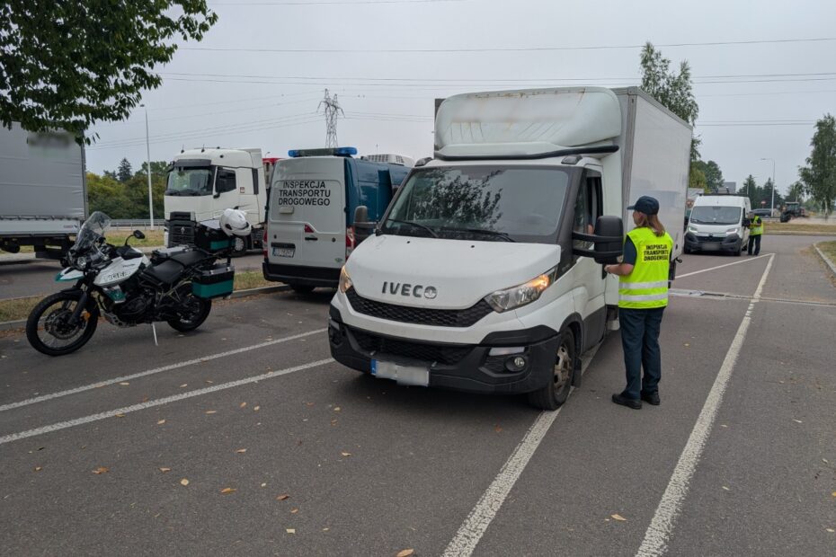 Kontrola drogowa prowadzona przez Inspekcję Transportu Drogowego; inspektor rozmawia z kierowcą dostawczego Iveco na parkingu obok radiowozu, motocykla służbowego i innych pojazdów.