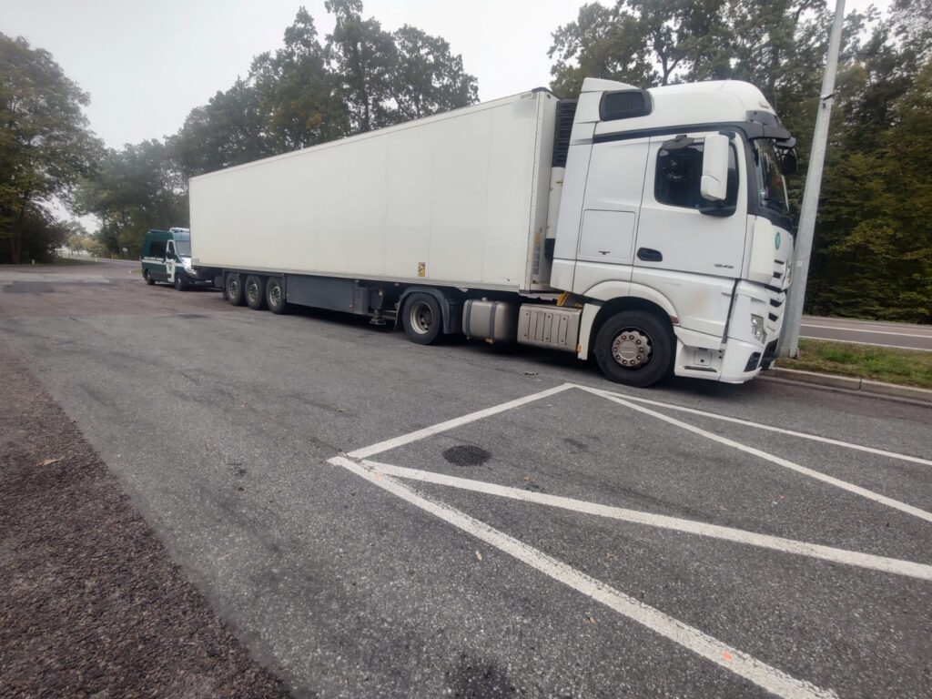 Biała ciężarówka z naczepą chłodnią zaparkowana na parkingu przy drodze.
