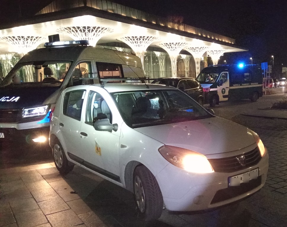 Taksówka zatrzymana do kontroli przez służby w centrum miasta nocą, w tle radiowozy policji i ITD.