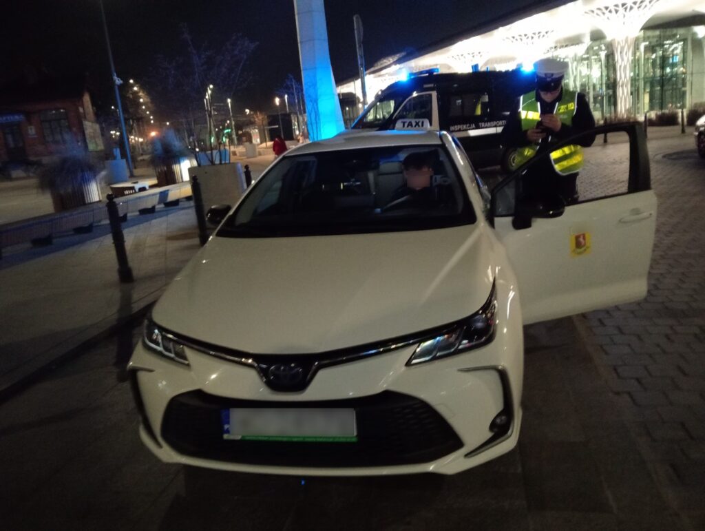Kontrola drogowa taksówki w nocy – funkcjonariusz sprawdza dokumenty kierowcy przy radiowozie z włączonymi światłami