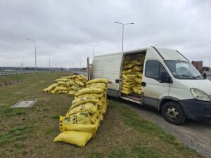 Biały samochód dostawczy z otwartą przestrzenią ładunkową, obok rozładowane worki z materiałem budowlanym przy drodze