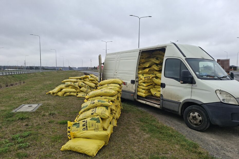 Biały samochód dostawczy z otwartą przestrzenią ładunkową, obok rozładowane worki z materiałem budowlanym przy drodze