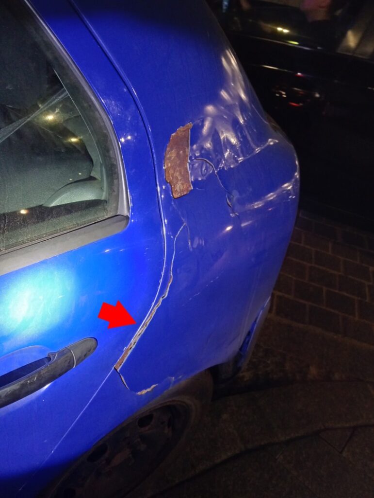 Uszkodzony niebieski samochód – wgnieciony i porysowany tylny błotnik z widoczną korozją