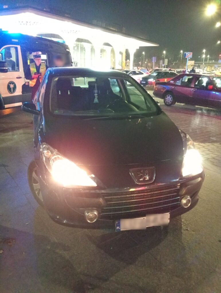 Czarny samochód osobowy zatrzymany do kontroli nocą przy dworcu, w tle radiowóz i inne pojazdy