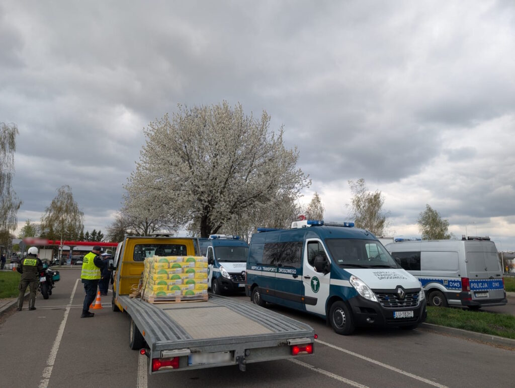 Laweta z ładunkiem materiałów budowlanych podczas kontroli drogowej, w tle pojazdy Inspekcji Transportu Drogowego i policji