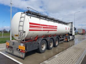 Biała naczepa cysterna z czerwonymi pasami, podpięta do ciągnika siodłowego Volvo, zaparkowana na betonowym placu postojowym przy drodze.